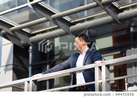 Asian businessman boss stands near his office center on the balcony in a dark color business suit Asian businessman boss stands near his office center on the balcony in a dark color business suit 94435939