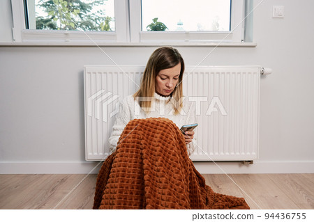 Worried sad woman sits under blanket near heating radiator and use smartphone, Rising costs in private households for gas bill due to inflation and war, Energy crisis Worried sad woman sits under blanket near heating radiator and use smartphone, Rising costs in private households for gas bill due to inflation and war, Energy crisis 94436755
