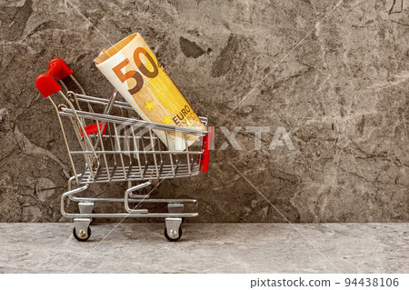 Shopping cart with roll of fifty Euro on the stone background Shopping cart with roll of fifty Euro on the stone background 94438106