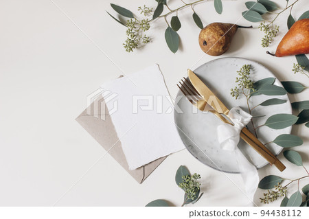 Autumn Thanksgiving table setting. Golden cutlery, plate, berry eucalyptus leaves, branches. Blank greeting card, invitation mockup. Pear fruit isolated on white table background. Fall flat lay, top. 94438112