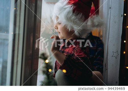 cute caucasian child wearing santa hat and plaid shirt sitting hugging by the window waiting for christmas 94439469