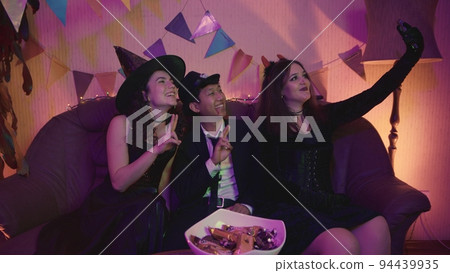 A group of friends are sitting at a table in creepy costumes, taking pictures on their phone and laughing on Halloween 94439935