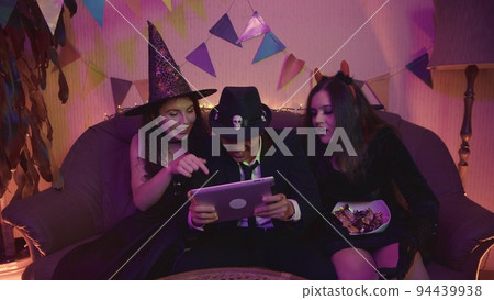 The guy in the creepy costume is actively playing on the tablet, and the girls cheerfully support him at the Halloween party. The guy in the creepy costume is actively playing on the tablet, and the girls cheerfully support him at the Halloween party. 94439938