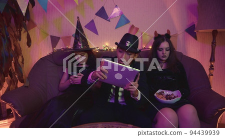 A guy in a creepy costume is actively playing on a tablet, and the girls are sitting on the phone at a Halloween party. 94439939