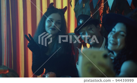 A group of friends are sitting at a table in creepy costumes, taking pictures with a camera and laughing on Halloween 94439999