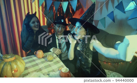 A group of friends are sitting at a table in creepy costumes, taking pictures with a camera and laughing on Halloween 94440000