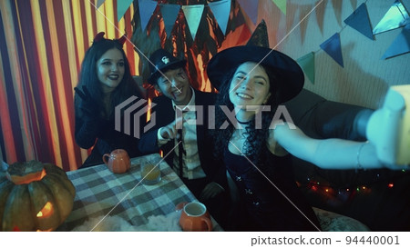 A group of friends are sitting at a table in creepy costumes, taking pictures with a camera and laughing on Halloween 94440001