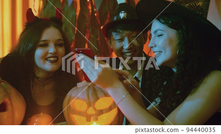 A group of friends are sitting at a table in creepy costumes and looking at the phone and laughing on Halloween 94440035