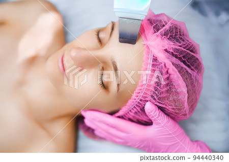 Beautician cosmetologist doing ultrasonic facial cleansing skin with device, professional cosmetic procedure in a beauty clinic salon for client. Skincare and machine cosmetology spa concept Beautician cosmetologist doing ultrasonic facial cleansing skin with device, professional cosmetic procedure in a beauty clinic salon for client. Skincare and machine cosmetology spa concept 94440340