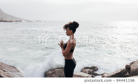 Side view of slim woman in sports clothes praying and meditating by ocean Side view of slim woman in sports clothes praying and meditating by ocean 94441196