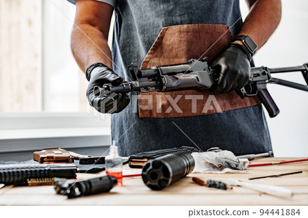 Close up of young man in apron disassembling a gun above the table Close up of young man in apron disassembling a gun above the table 94441884
