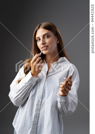 Cute young woman applying lip gloss against gray background Cute young woman applying lip gloss against gray background 94441928