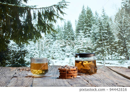 Glass pot with herbal tea, cakes, biscuits on wooden table over winter snow forest background. Design for greeting card. Holidays, vacation, traditions Glass pot with herbal tea, cakes, biscuits on wooden table over winter snow forest background. Design for greeting card. Holidays, vacation, traditions 94442816