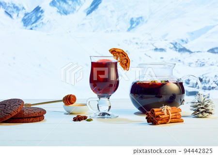 Hot mulled wine in glasses with orange slices, cinnamon sticks and spices with Christmas decorations on table over background of landscape of winter snow-capped mountains 94442825