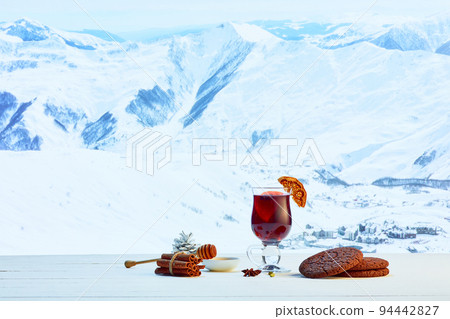 Design for greeting card. Glass with mulled wine, cakes, biscuits, honey and Christmas decor on table over winter snow-capped mountains background Design for greeting card. Glass with mulled wine, cakes, biscuits, honey and Christmas decor on table over winter snow-capped mountains background 94442827