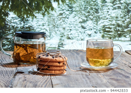Winter still life. Glass pot with herbal tea, cakes, biscuits on wooden table over winter snow forest background. Design for greeting card, wallpaper for device screen Winter still life. Glass pot with herbal tea, cakes, biscuits on wooden table over winter snow forest background. Design for greeting card, wallpaper for device screen 94442833