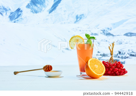 Glass with orange tea or cocktail, berries and honey on white table over background of landscape of winter snow-capped mountains. Health, care, love Glass with orange tea or cocktail, berries and honey on white table over background of landscape of winter snow-capped mountains. Health, care, love 94442918