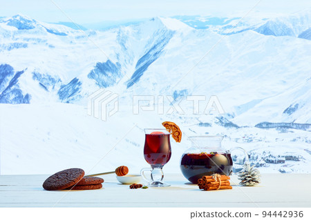 Hot mulled wine in glasses with orange slices, cinnamon sticks and spices with Christmas decorations on table over background of landscape of winter snow-capped mountains 94442936