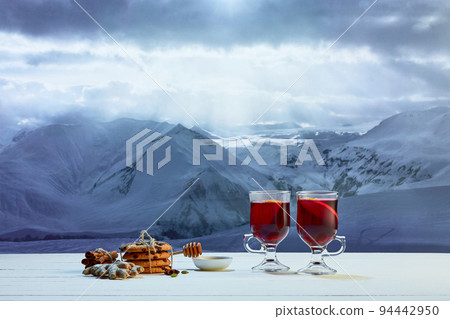 Design for greeting card. Glass with mulled wine, cakes, biscuits, honey and Christmas decor on table over winter snow-capped mountains background Design for greeting card. Glass with mulled wine, cakes, biscuits, honey and Christmas decor on table over winter snow-capped mountains background 94442950
