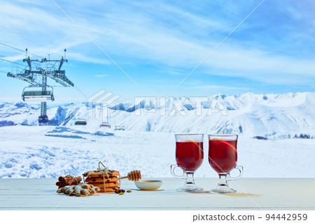 Hot mulled wine in glasses with orange slices, cinnamon sticks and spices with Christmas decorations on table over background of landscape of winter snow-capped mountains Hot mulled wine in glasses with orange slices, cinnamon sticks and spices with Christmas decorations on table over background of landscape of winter snow-capped mountains 94442959