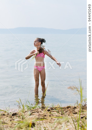 Teenage girl in a swimsuit fanning herself with a broom of grass imitating bath procedures on the sunny river bank in summer Teenage girl in a swimsuit fanning herself with a broom of grass imitating bath procedures on the sunny river bank in summer 94445049
