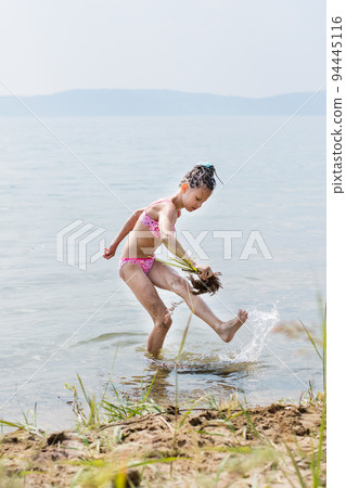 A teenage girl in a bathing suit fanns her leg with a broom of grass imitating bath procedures on the sunny bank of the river in summer. Vacation activities for children 94445116