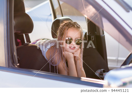 Smiling teenage girl with afro-braids sits in the passenger compartment of a car on a hot summer day. Road trip 94445121