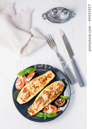 Ready-to-eat baked zucchini halves filled with cheese and tomato and basil leaves and cutlery on a black plate on a white table. Vegetable menu, healthy food. Top and vertical view 94445957