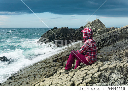 traveler sits on the shore of a stormy sea, on a natural pavement made of columnar granite traveler sits on the shore of a stormy sea, on a natural pavement made of columnar granite 94446316