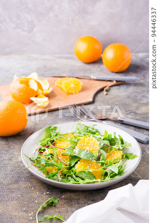 Diet vegetarian vitamin salad of orange slices and a mix of arugula, chard and mizun leaves on a plate and a cutting board with peeled orange on the table. Vertical view 94446337