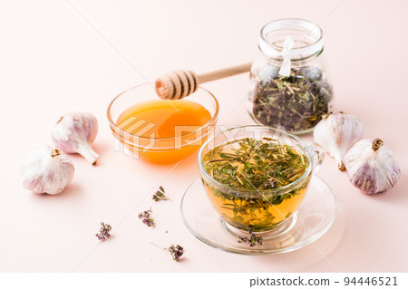 Hot tea with oregano in a cup, honey in a bowl, heads of garlic and dry herb in a jar. Herbal medicine and alternative therapy 94446521