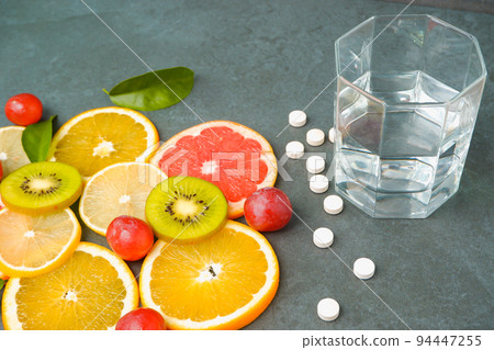 Supplements or healthy natural food? Scattered white pills vitamins, Sliced citrus fruits, orange, lemon, kiwi, grapes, glass with water on a stone gray background Supplements or healthy natural food? Scattered white pills vitamins, Sliced citrus fruits, orange, lemon, kiwi, grapes, glass with water on a stone gray background 94447255