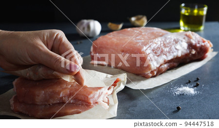 Cooking food. A woman's hand wraps chopped steaks in paper. Fresh raw meat with spices against a dark background. 94447518