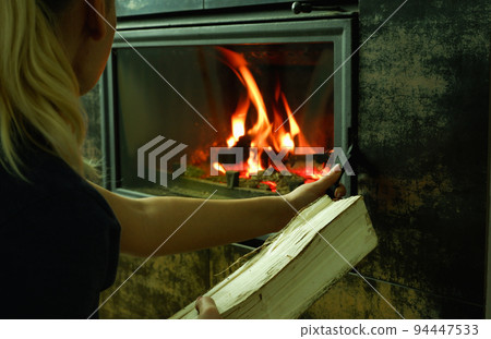 A woman's hand puts wood in the burning fireplace. Modern closed fireplace with glass. 94447533
