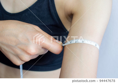 Close up of young woman in sport bra using a measuring tape for measuring her arm length. 94447678