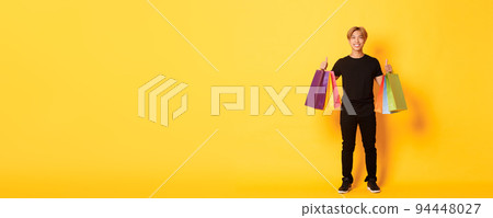 Full-length of attractive korean guy in black clothes, showing thumbs-up, holding shopping bags, yellow background Full-length of attractive korean guy in black clothes, showing thumbs-up, holding shopping bags, yellow background 94448027