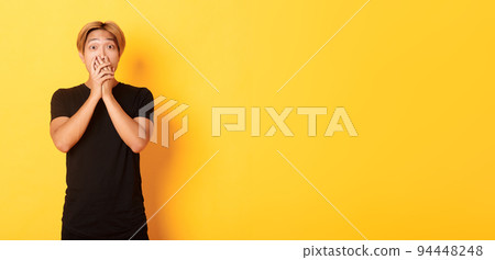 Portrait of surprised asian handsome guy with blond hair, cover mouth with hands and stare at camera impressed, yellow background 94448248