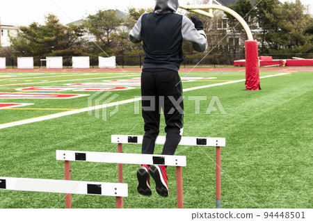 Athlete in fulll sweatsuit jumping over track hurdles on a turf field Athlete in fulll sweatsuit jumping over track hurdles on a turf field 94448501