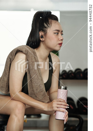 Exhausted young sportswoman resting on bench and drinking fresh water after working out in gym Exhausted young sportswoman resting on bench and drinking fresh water after working out in gym 94448782