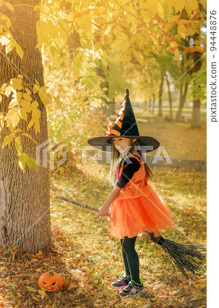 A girl in the guise of a witch sits astride a broomstick in the forest between the autumn yellow foliage of trees on Halloween 94448876