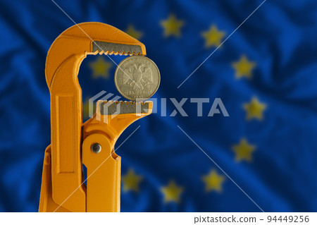 Russian two rubles coin clamped in an orange plumber wrench on the background of the European Flag. Russian two rubles coin clamped in an orange plumber wrench on the background of the European Flag. 94449256