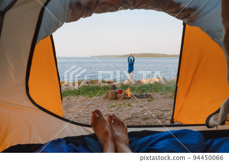 view from a tent on the shore of a lake or sea, a woman collects firewood or does yoga and meditation. camping in nature view from a tent on the shore of a lake or sea, a woman collects firewood or does yoga and meditation. camping in nature 94450066