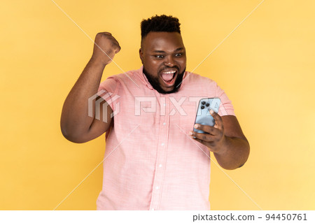 Portrait of satisfied bearded man wearing pink shirt standing with cell phone in hands and clenching fist, celebrating his victory, looks at screen. Indoor studio shot isolated on yellow background. 94450761