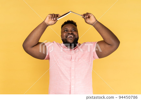 Portrait of joyful delighted bearded man wearing pink shirt holding opened book on head, looking up with happy positive expression. Indoor studio shot isolated on yellow background. 94450786