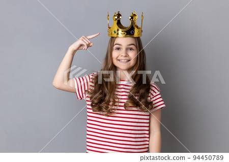 Portrait of little girl princess wearing striped T-shirt pointing fingers on golden crown on her head, showing her leadership qualities. Indoor studio shot isolated on gray background. 94450789