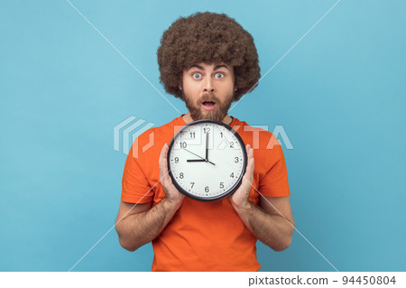 Portrait of shocked man with Afro hairstyle being late, holding in hand wall watch, deadline, punctuality, looking at camera with open mouth. Indoor studio shot isolated on blue background. 94450804
