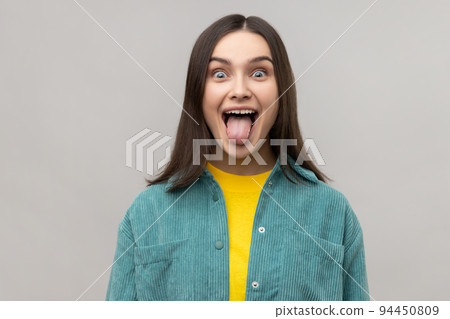 Dark haired woman sticking out tongue and looking at camera, teasing with naughty expression, disobedient behavior, wearing casual style jacket. Indoor studio shot isolated on gray background. Dark haired woman sticking out tongue and looking at camera, teasing with naughty expression, disobedient behavior, wearing casual style jacket. Indoor studio shot isolated on gray background. 94450809