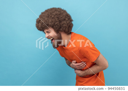 Side view of laughing man with Afro hairstyle in orange T-shirt holding his stomach and hunched in crazy hysterical laughter, sincere joyful emotions. Indoor studio shot isolated on blue background. 94450831