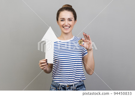 Portrait of positive attractive woman wearing striped T-shirt showing gold bitcoin and arrow pointing up, growth of electronic currency . Indoor studio shot isolated on gray background. 94450833