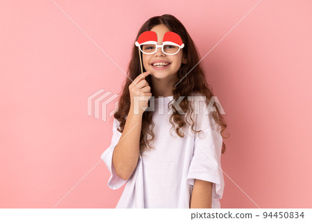 Portrait of little girl wearing white T-shirt holding funny paper glasses, having fun playing game, wearing masquerade accessory. Indoor studio shot isolated on pink background. Portrait of little girl wearing white T-shirt holding funny paper glasses, having fun playing game, wearing masquerade accessory. Indoor studio shot isolated on pink background. 94450834